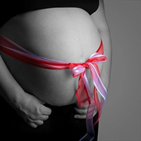 Ein Babybauch, stilvoll umwickelt mit einer roten und pinkfarbenen Schleife, die zu einer Schleife gebunden ist. Das Bild ist größtenteils Schwarz-Weiß, wobei die Schleife farbig hervorsticht. Ein symbolträchtiges Porträt der Vorfreude auf das "Geschenk" des Lebens, festgehalten von Fotograf Michael Schalansky.